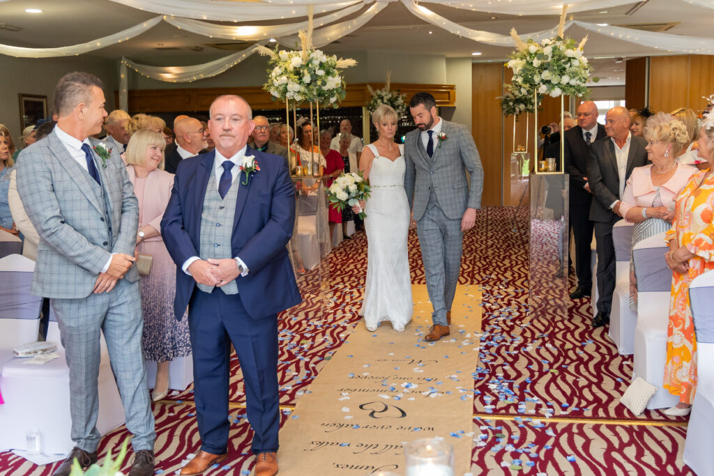 A bride in a white dress walks down the aisle with a man in a grey suit, approaching a man in a blue suit at an indoor wedding ceremony with seated guests. Focus Photography by Tim Teasdale