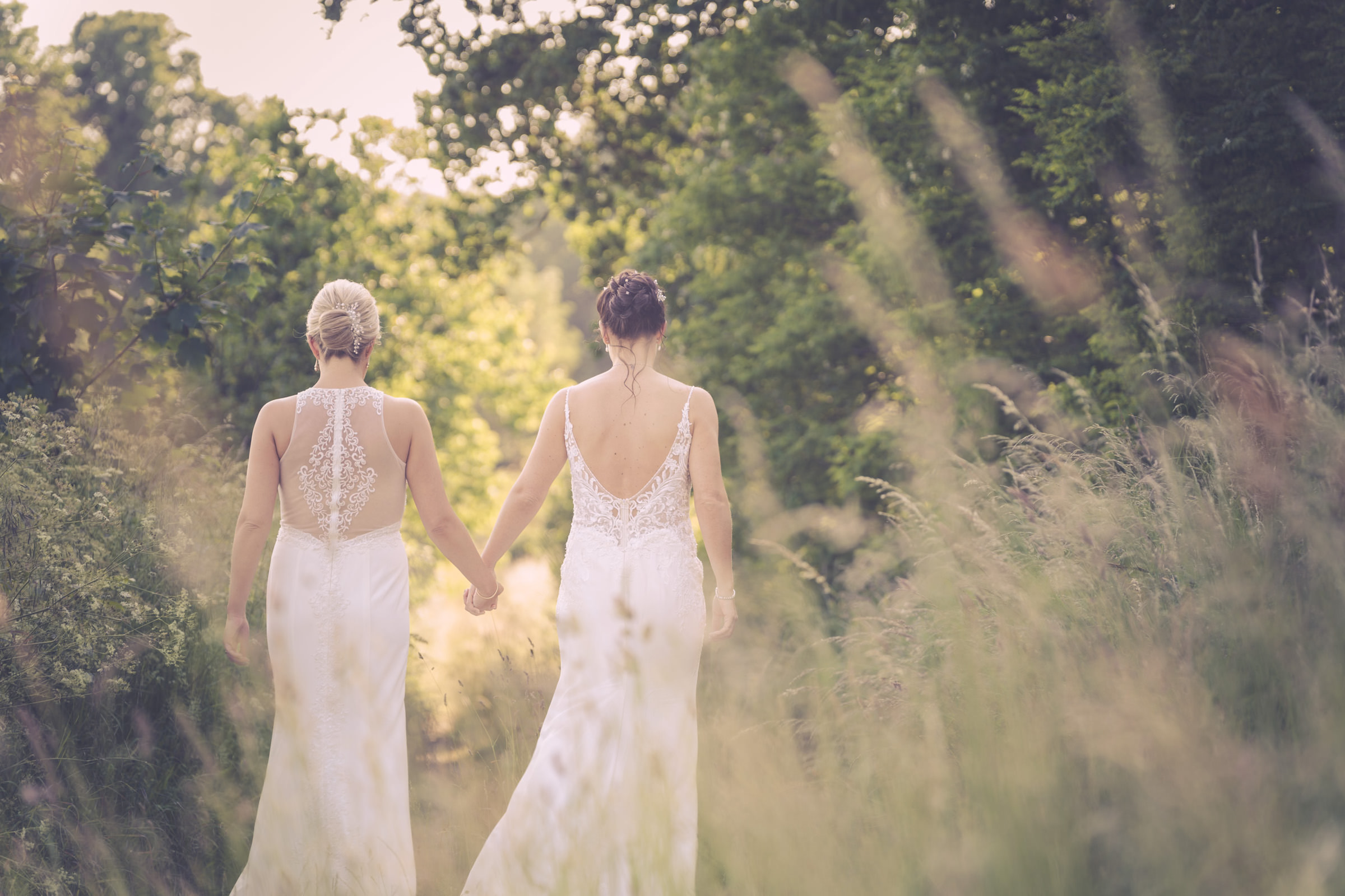 Two women in white wedding dresses walk hand in hand along a grassy outdoor path, surrounded by greenery and soft sunlight