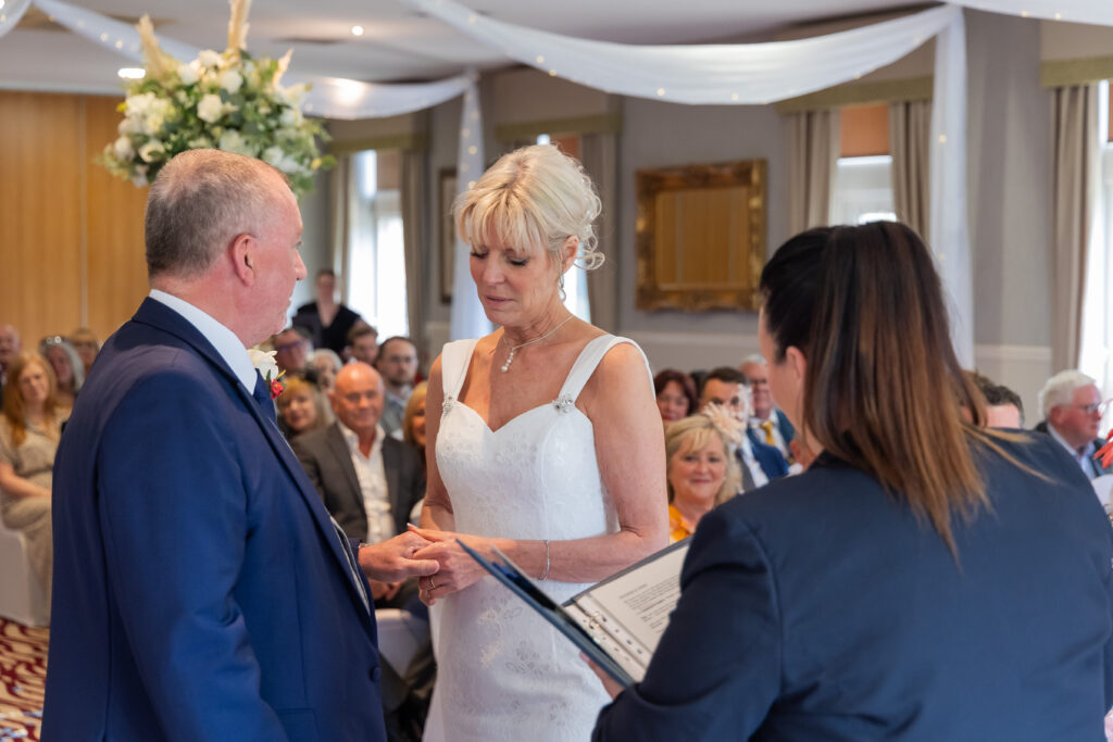 A bride and groom stand facing each other during their wedding ceremony, exchanging rings with an officiant and guests seated in the background. Focus Photography by Tim Teasdale