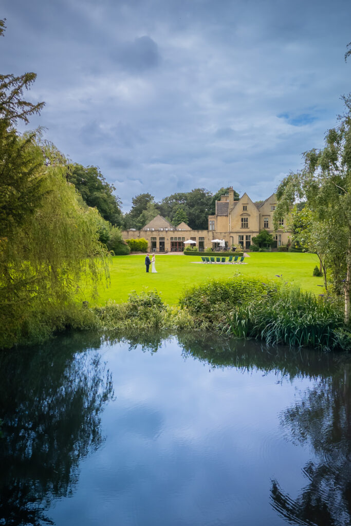 A couple stands on a large green lawn in front of a manor house, with a pond in the foreground and trees surrounding the scene under a cloudy sky. Focus Photography by Tim Teasdale