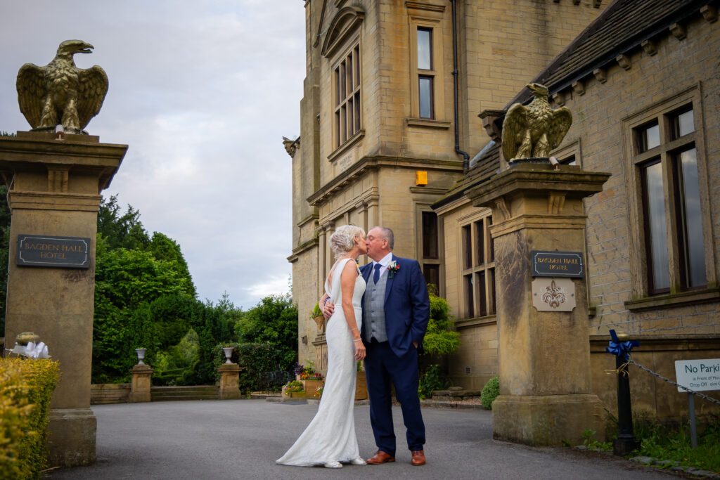 A bride and groom share a kiss outside the entrance of Bagden Hall Hotel, flanked by stone pillars topped with eagle statues. Focus Photography by Tim Teasdale