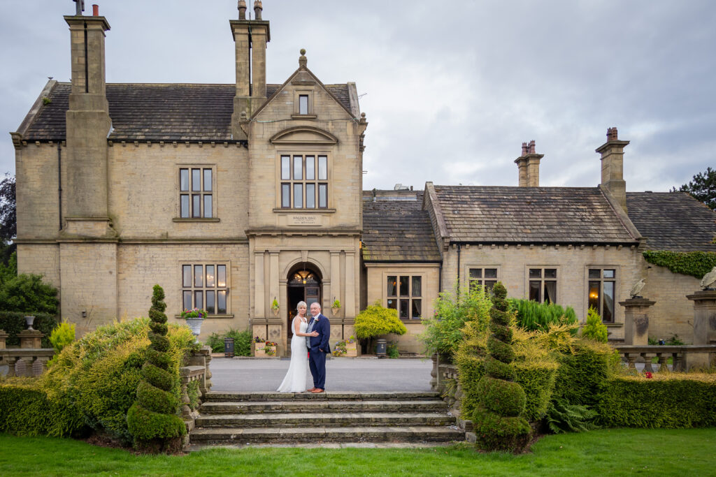 A couple in formal attire stands on stone steps in front of a large, historic stone manor with manicured gardens. Focus Photography by Tim Teasdale