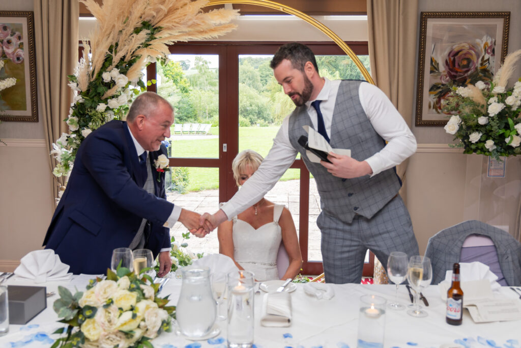 A man in a grey suit shakes hands with an older man in a navy suit at a wedding reception table, whilst a woman in a white dress sits between them. Focus Photography by Tim Teasdale