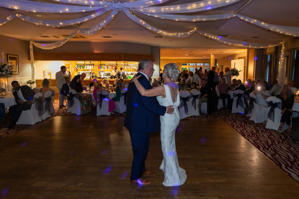 A bride and groom share their first dance in a decorated reception hall with seated guests watching. Focus Photography by Tim Teasdale
