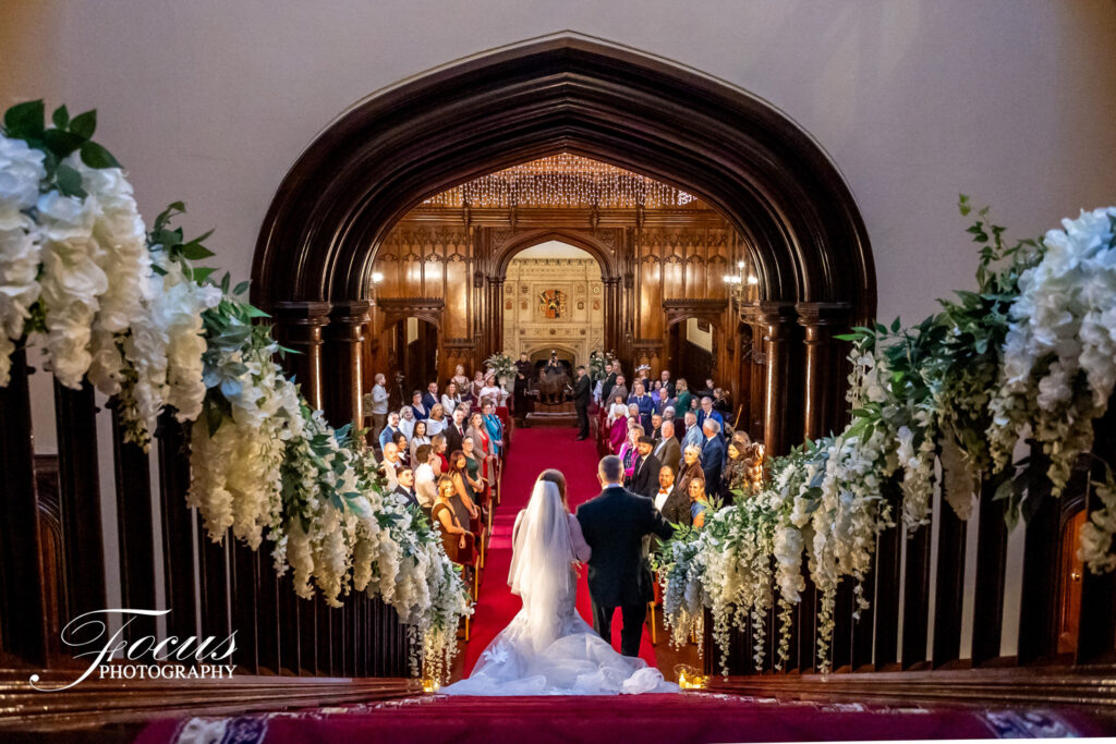 A bride and groom stand at the top of a flower-adorned staircase, facing guests seated in a grand hall decorated for a wedding ceremony. Focus Photography by Tim Teasdale