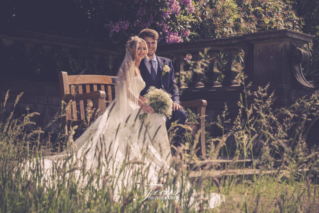 A bride and groom sit together on a wooden bench outdoors, surrounded by greenery and flowers, with sunlight filtering through the trees
