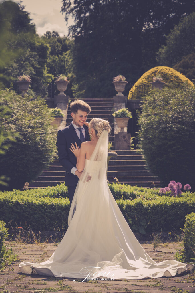 A bride and groom stand facing each other in a garden, smiling, with steps and greenery in the background. The bride wears a white dress and veil; the groom is in a dark suit