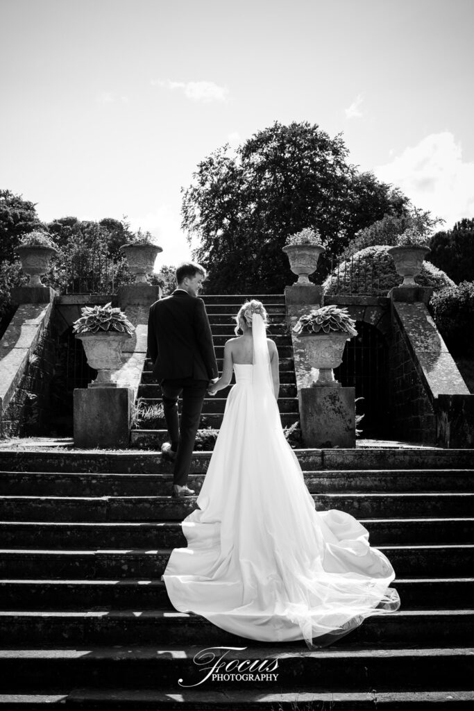 A bride and groom walk up outdoor stone steps, holding hands, with the bride in a long white dress and the groom in a suit