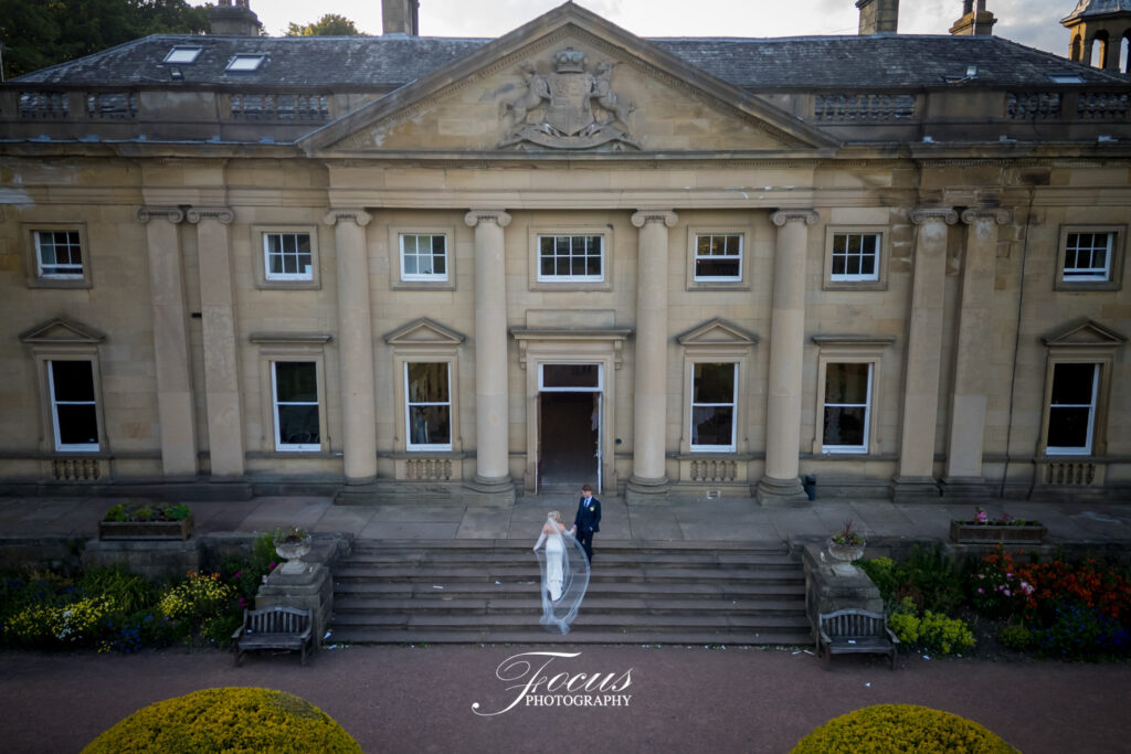 A bride and groom stand on the steps outside a large, neoclassical building with columns and landscaped gardens