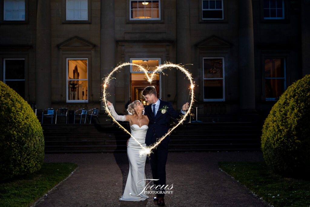 A bride and groom in formal attire create a heart shape with sparklers outside a large building at night
