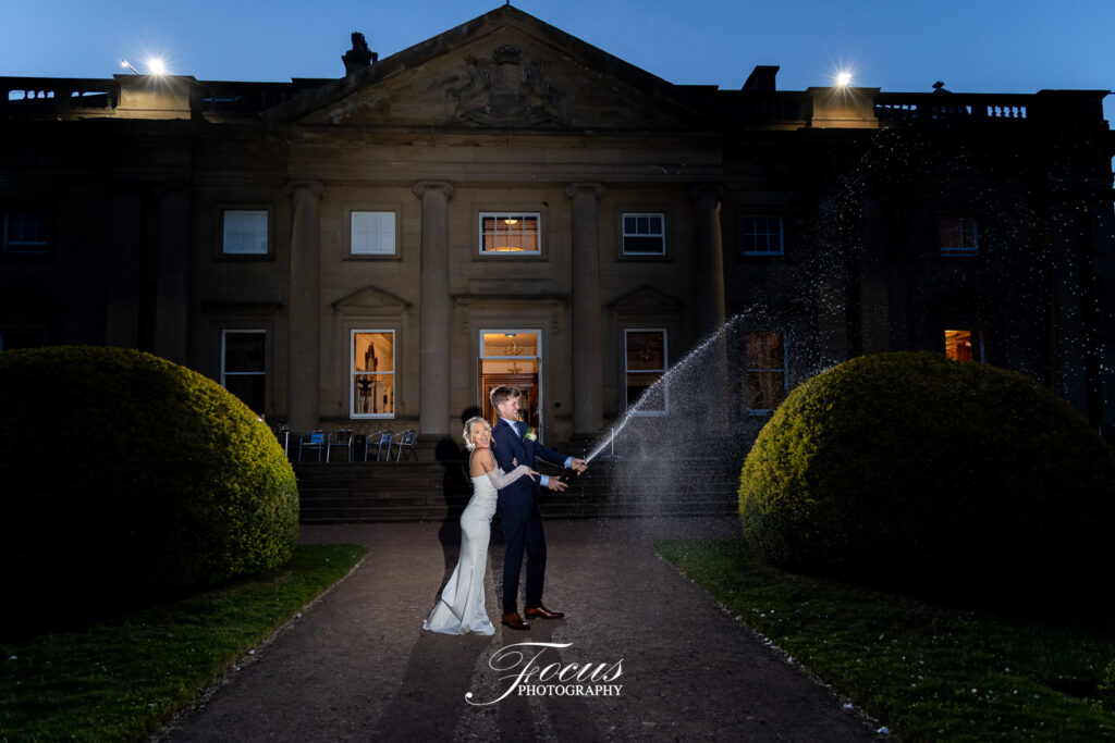 A bride and groom stand on a path at night in front of a large building, opening a bottle of champagne with the spray captured in mid-air